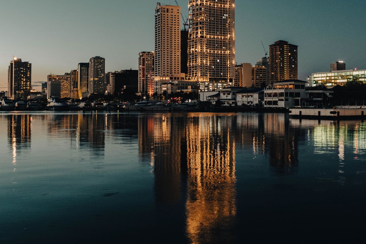A waterfront skyline at twilight, with a tall tower outlined in warm lights reflected in calm water.