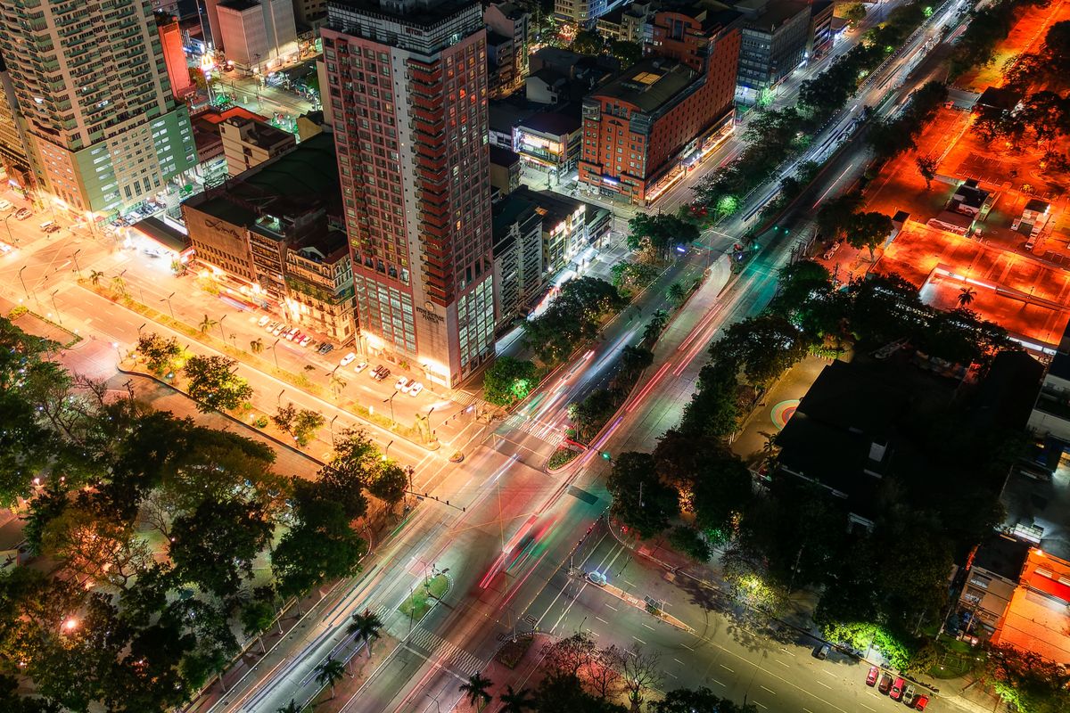 A steep aerial view at night of a big intersection with palm-lined boulevards, light trails and lit high-rises.