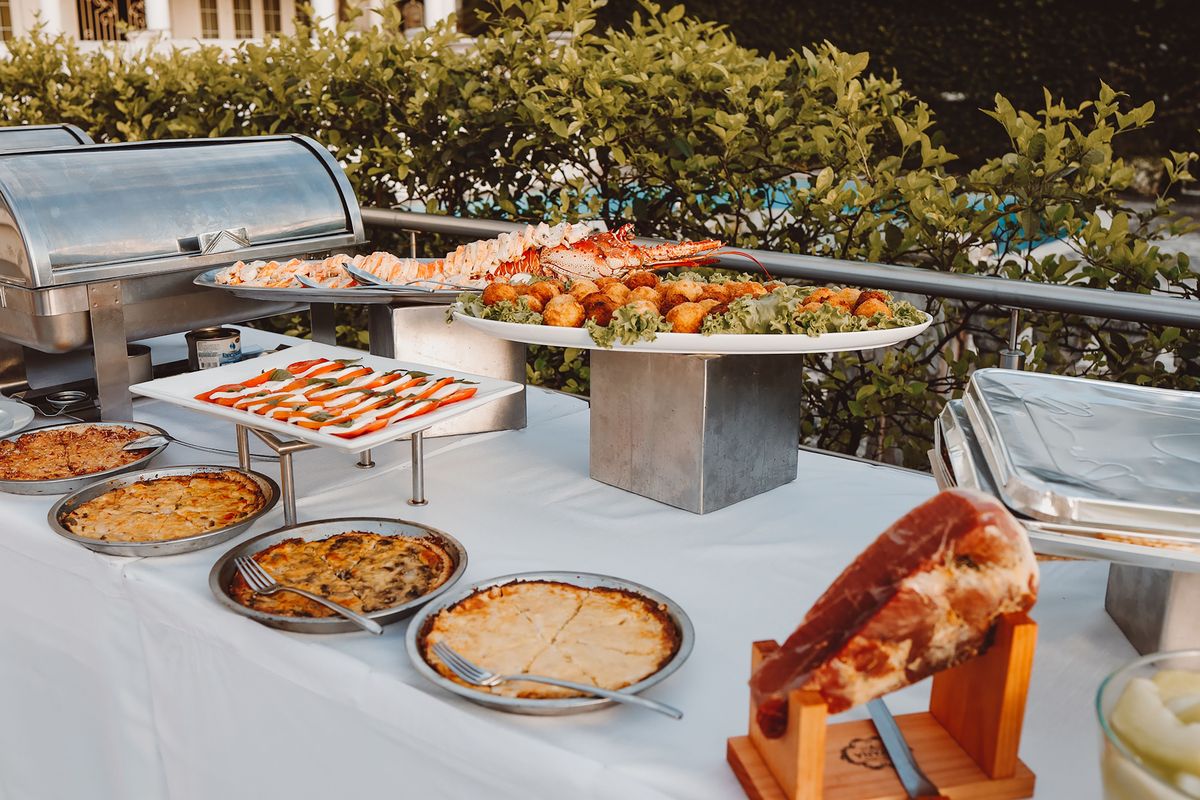 A buffet table set outdoors with chafing dishes, quiches, salads, a platter of seafood and a cured ham on its stand.