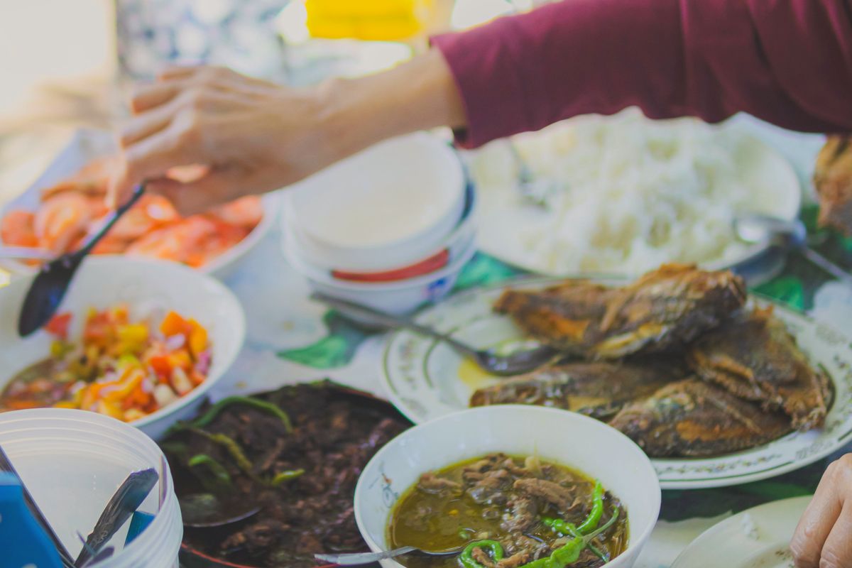 Hands reaching across a family table of fried fish, rice, ensalada and soup with chilies.