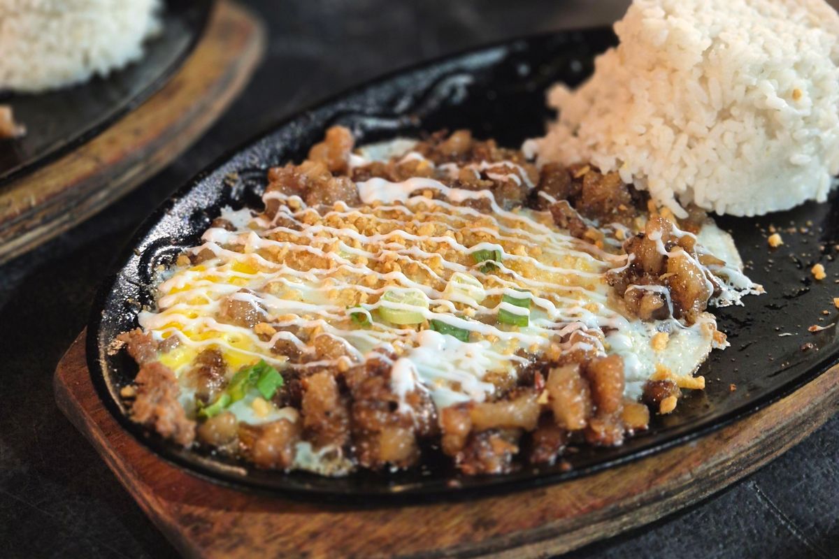 Pork sisig topped with mayonnaise and spring onion on a cast-iron sizzling plate, next to a dome of white rice.