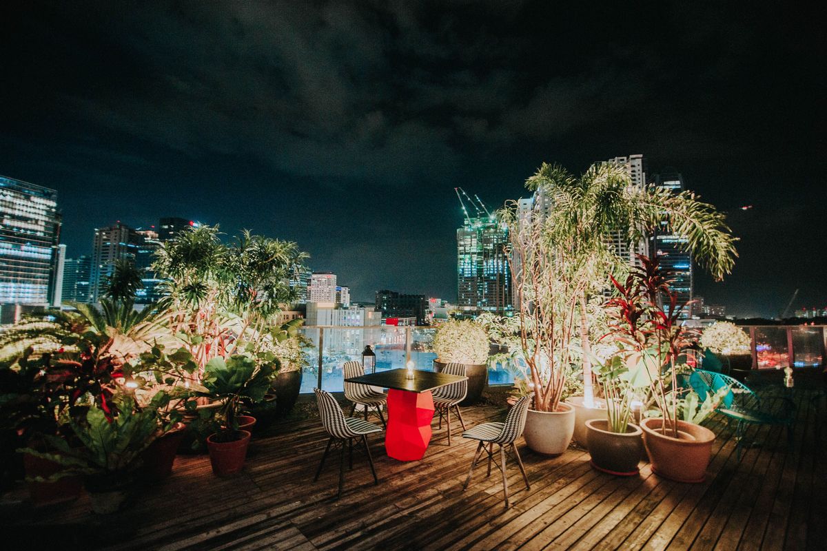 A rooftop terrace at night with potted palms, a candle-lit table and lit towers behind.