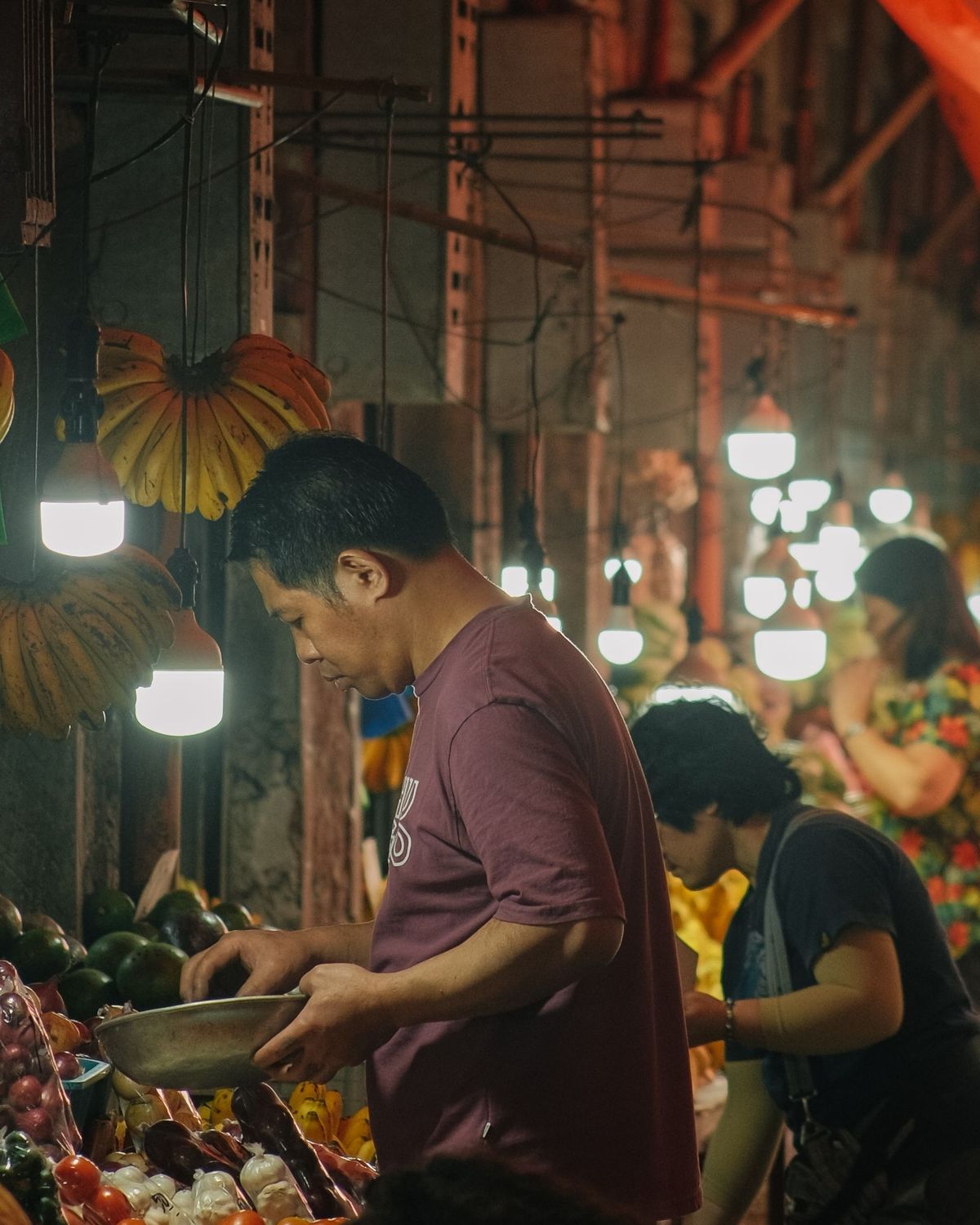 A vendor weighing produce under hanging bulbs in a night-market aisle.