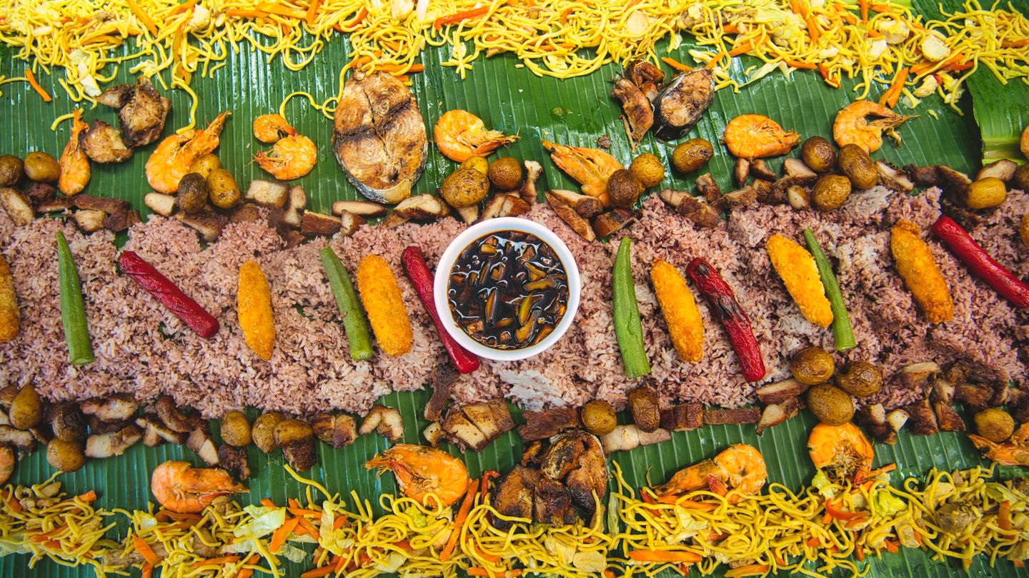 A boodle fight laid out on banana leaves, seen from above: rice, grilled fish, shrimp and noodles around a bowl of soy-vinegar dip.