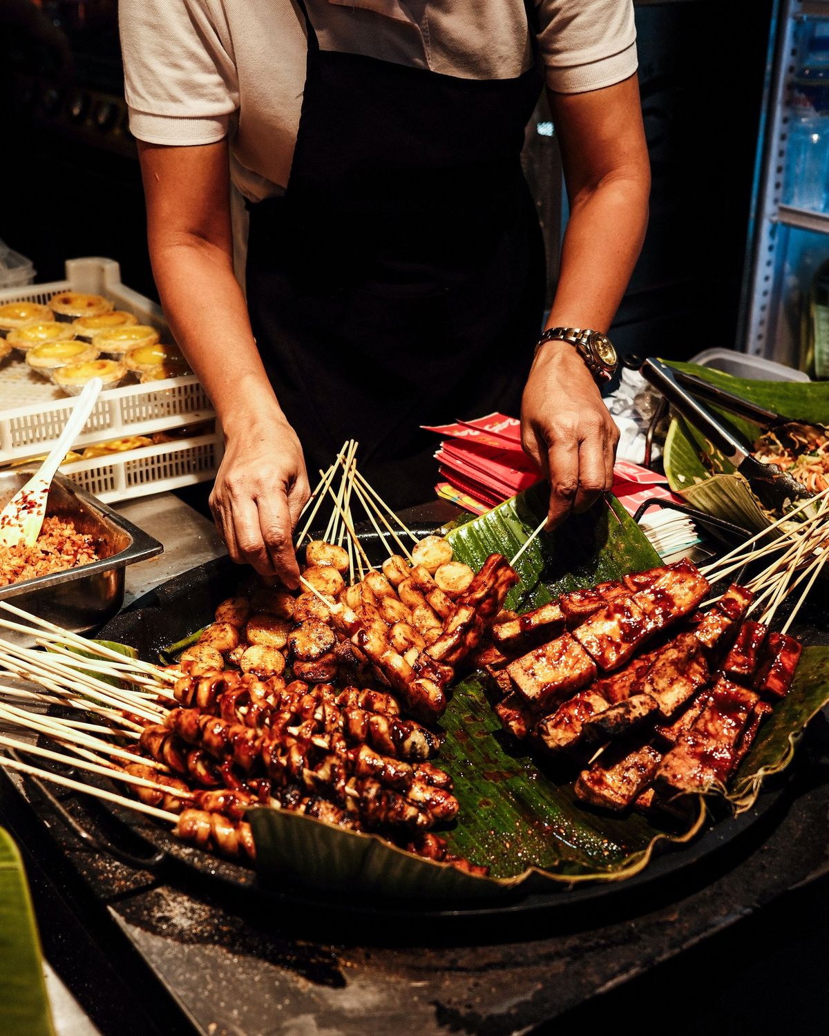 A street-food vendor arranging glazed barbecue skewers on banana leaves over a griddle at night.