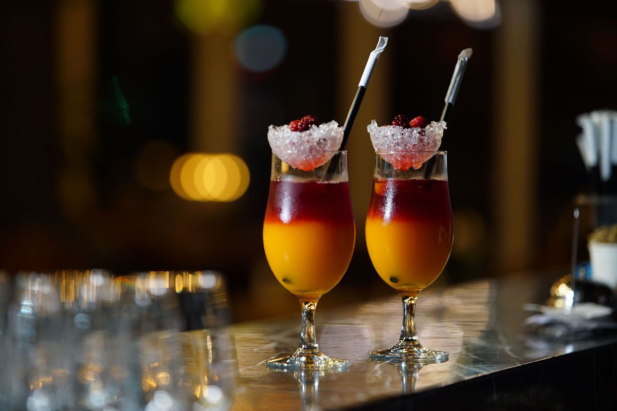 Two layered red and orange cocktails topped with crushed ice and berries on a dark stone bar counter, with soft lights blurred behind them.