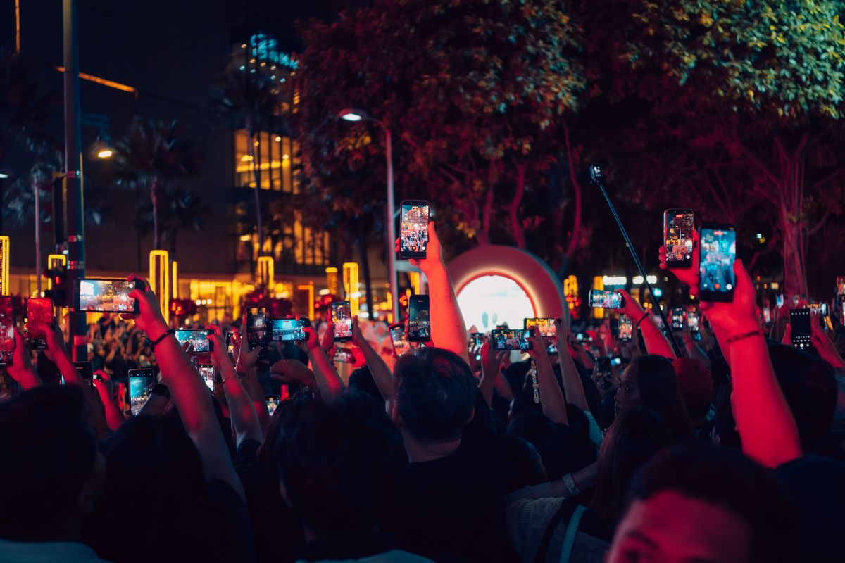 A night crowd seen from behind, holding up glowing phones to film an event.