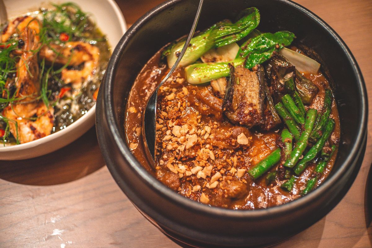 A dark clay pot of kare-kare, meat stew in a thick peanut sauce topped with crushed peanuts, bok choy, eggplant and long beans, next to a bowl of shrimp, on a wooden table.
