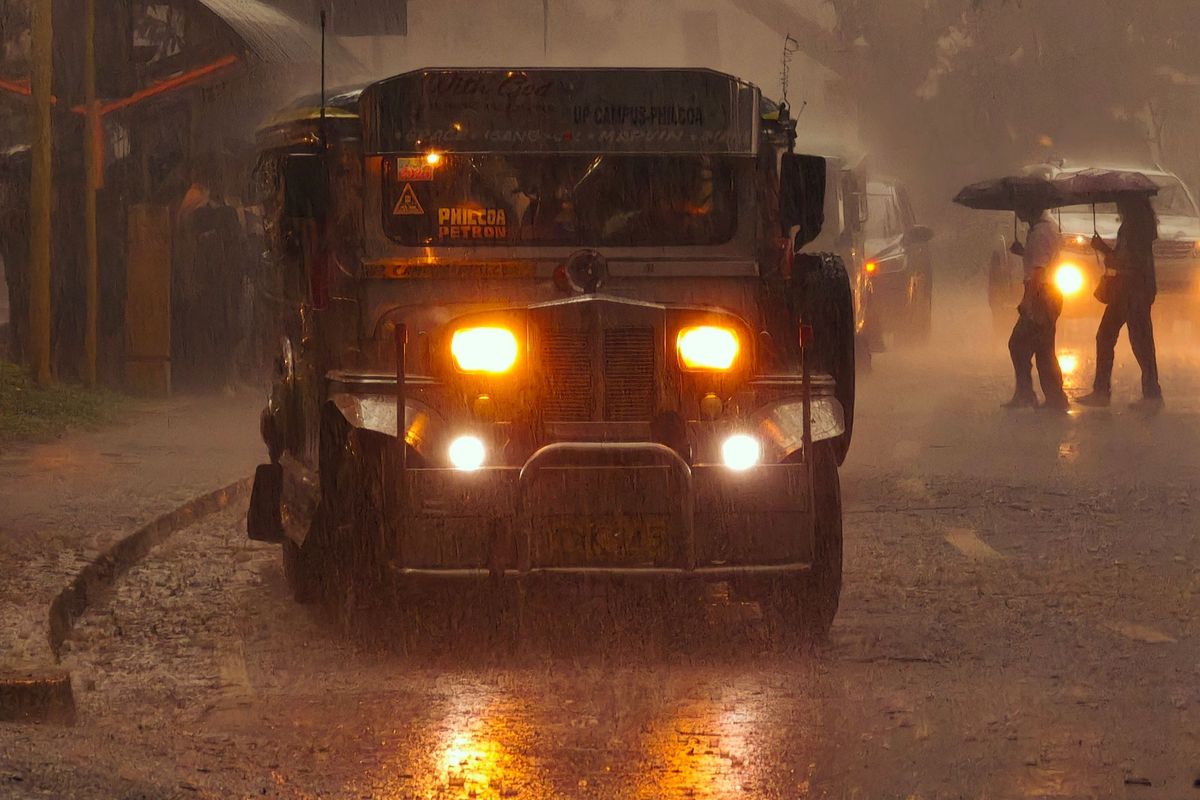 A jeepney drives head-on through heavy rain with its headlights glowing, reflections streaming across the wet road.