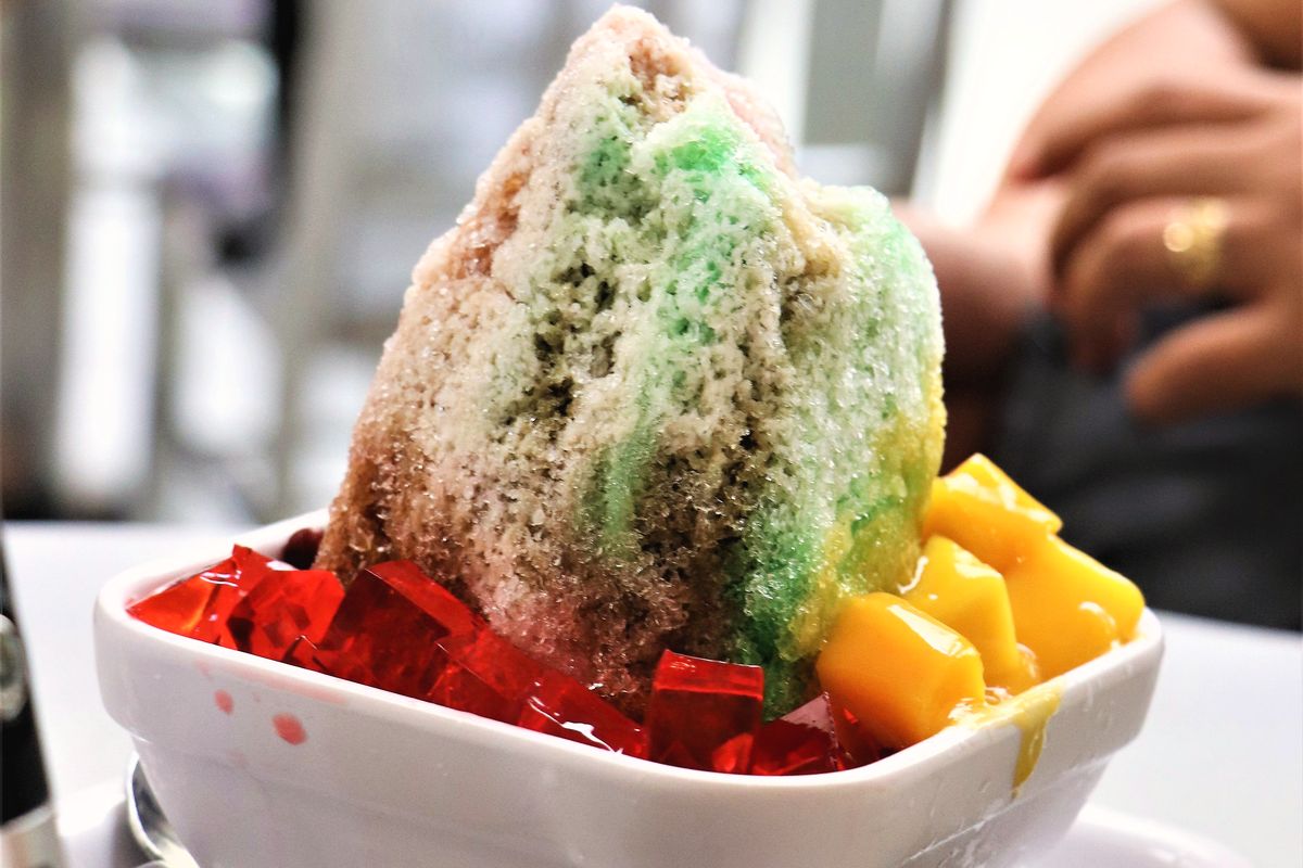 A tall peak of tinted shaved ice in a white bowl, with red gelatin cubes and diced mango at its base.