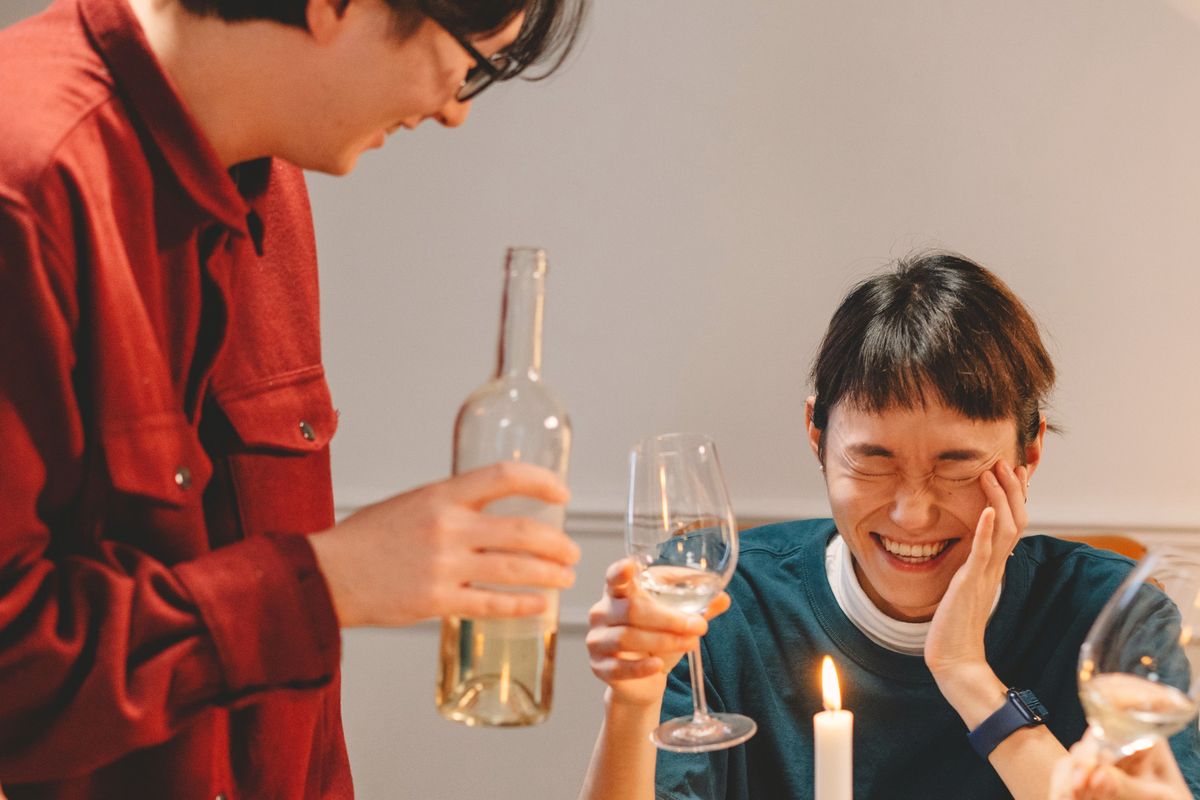 Two friends at a candlelit dinner table: one stands holding a wine bottle, the other laughs with a hand on her cheek.