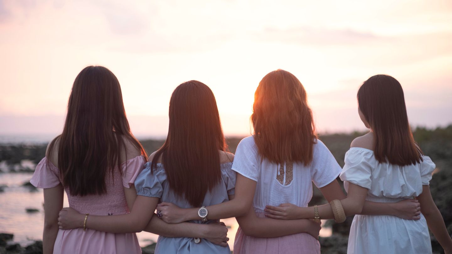 Four young women with their arms around each other, seen from behind, facing a pink sunset over a rocky shore.