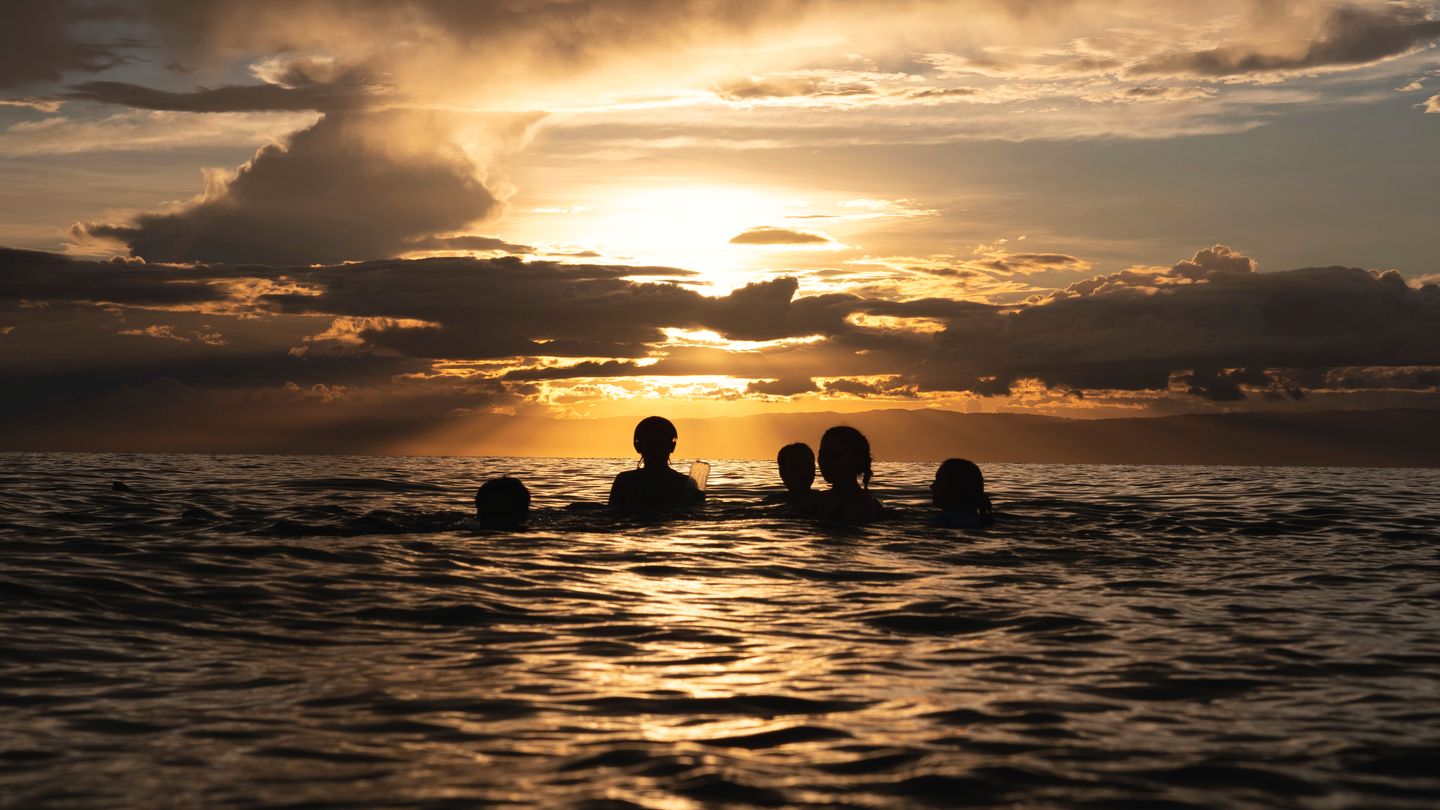 Five people swimming in the sea, in silhouette against a golden sunset.