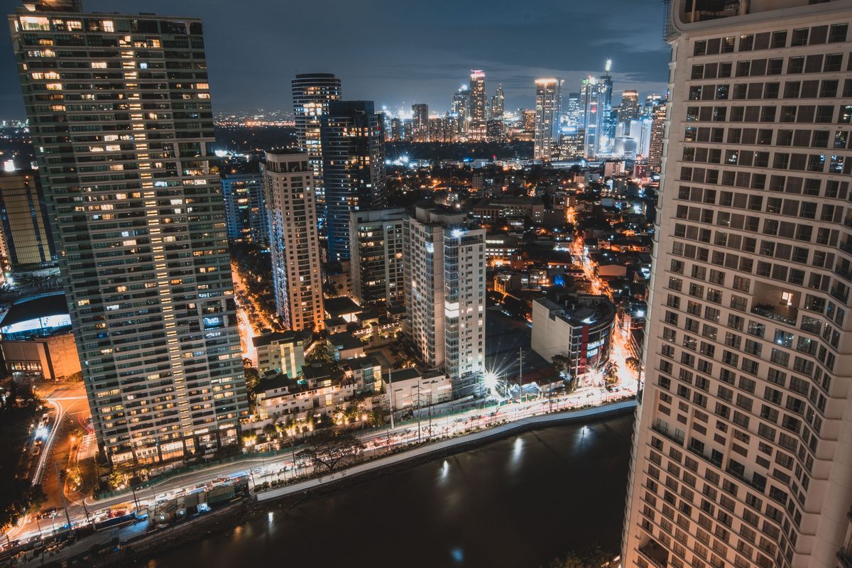 Looking down from a high-rise at night: a dark river bends between lit towers, with a skyline on the horizon.