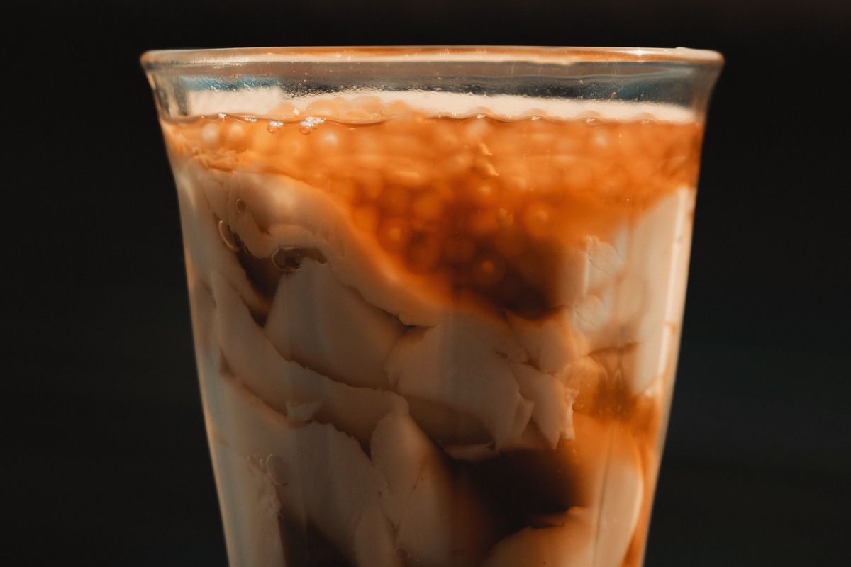 A glass of taho: silken tofu swirled with dark arnibal syrup and sago pearls, in warm light against a dark background.